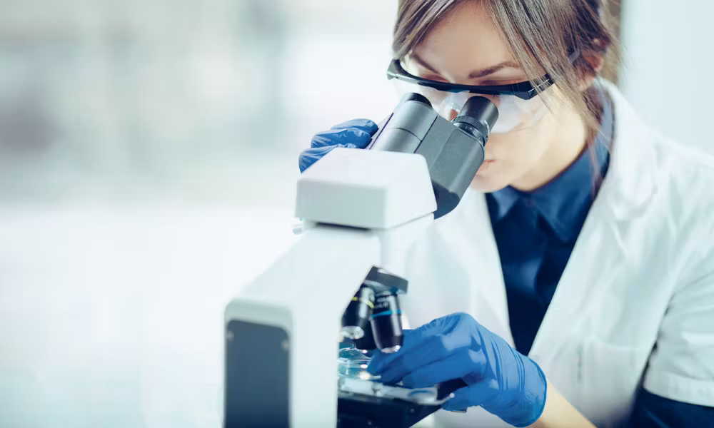 Une scientifique portant des gants et des lunettes de protection observe un échantillon à travers un microscope dans un laboratoire.